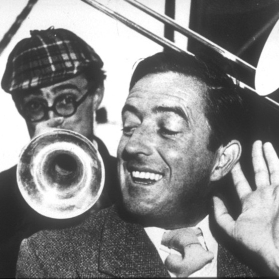 Black and white photo of a close up of a man with his left hand raised up to his ear. Over his shoulder is a man with glasses and a tartan pattern cap, man has rested the end of a trumpet.