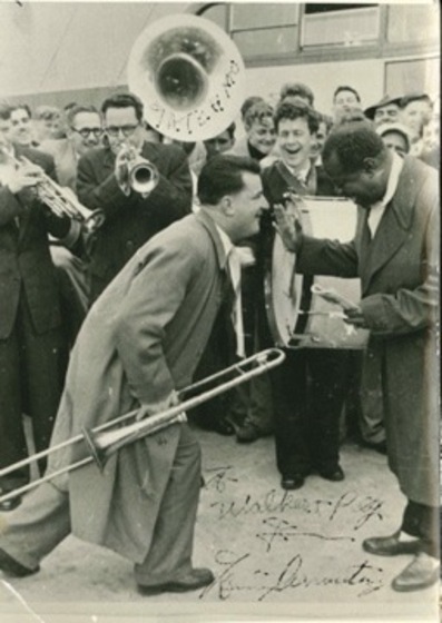Black and white photo of a man holding a trombone and stepping towards a man in front of him. Behind is a crowd of people, including some who hold drums and trumpets.