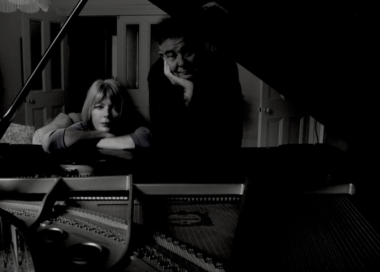 Black and white photo of a woman and a man leaning on an open grand piano cast in shadow. They are standing where the keys are, with the view taken from the other side of the piano.