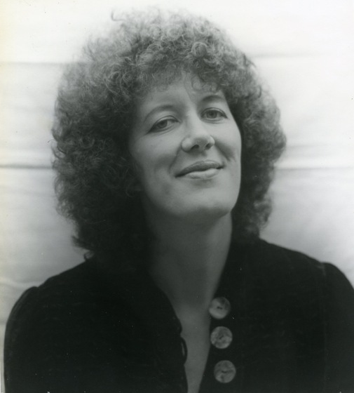 Black and white photo of headshot of a woman with curly hair, wearing a black top with three large buttons at the neck.