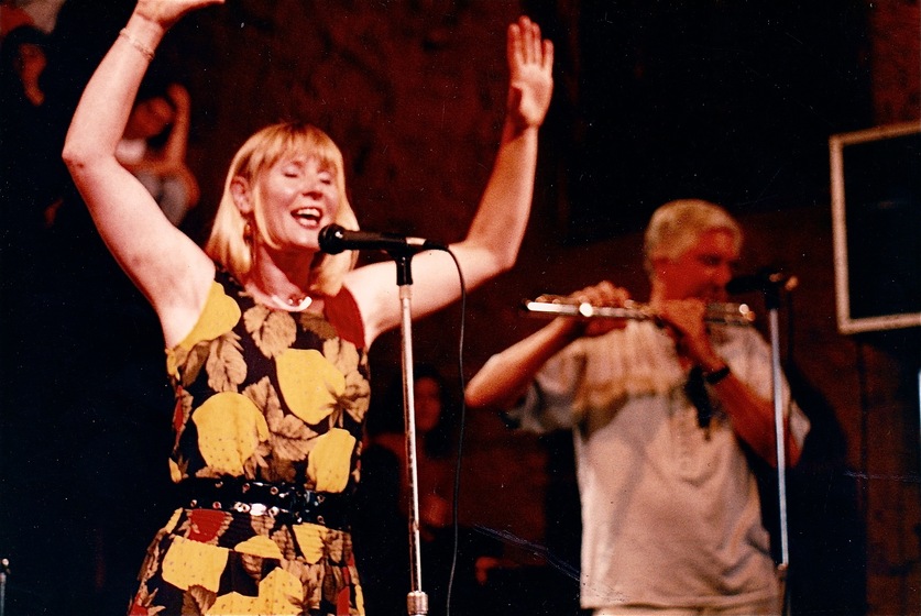 Colour photo of a woman with a yellow leaf patterned dress standing in front of a microphone with hands raised. Behind her out of focus is a man playing a flute behind a microphone.  