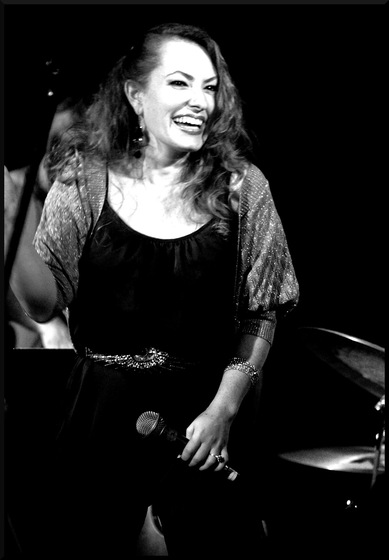 Black and white photo of a woman in a black top and pants, with a lighter coloured short sleeved jacket, holding a microphone down low in front of her with a big smile on her face. 
