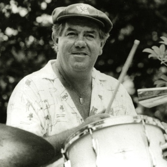 Black and white photo of a man seated behind a drum kit holding up one drumstick. He wears a cap and a patterned light shirt, and shrubs can be seen behind him.