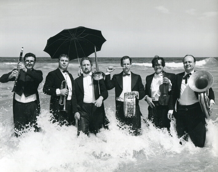 Black and white photo of six men wearing three-piece suits with tails standing in the ocean as a wave crashes up against the back of their legs. From left to right they hold, a clarinet, a trumpet and umbrella, a trombone, a small drum, a violin, and a tuba.