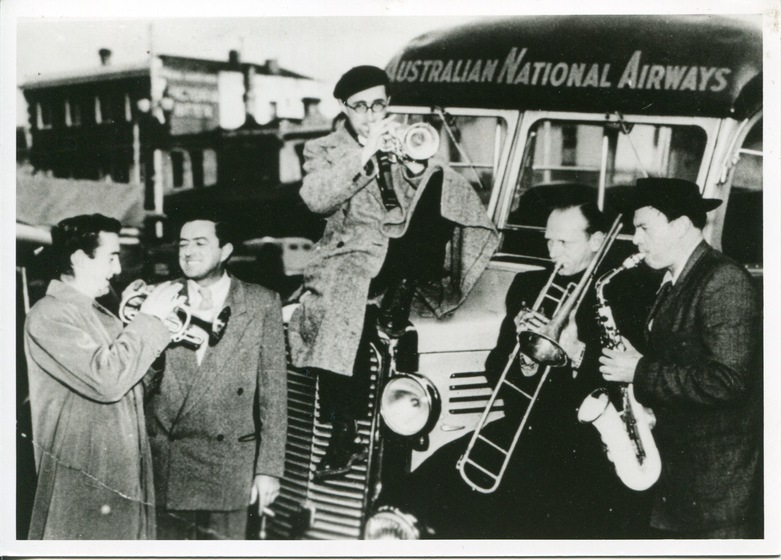 Black and white photograph of five men playing instruments in front of a bus with 'Australian National Airways' on the front. The man on the left plays the cornet, the left man from the left has no instrument. The man in the centre sits on the bus hood playing a trumpet. To the right is a man playing the trombone, and a man playing a saxophone.