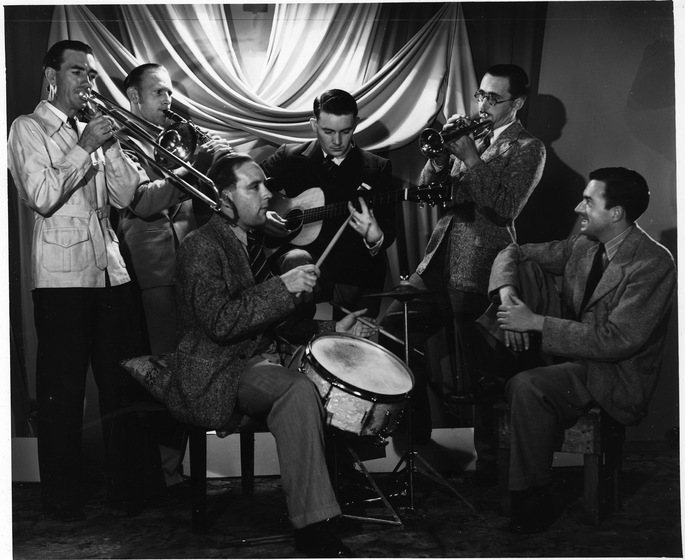 Black and white photograph of six men seated and standing playing instruments with draped fabric behind. They play from left to right, a trombone, clarinet, drum, guitar, and trumpet. One man is seated on the right with no intrument.