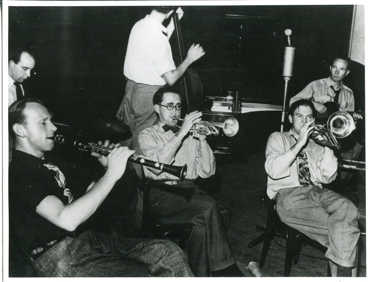 Black and white photograph of three men seated playing instruments. From left to right they play a clarinet, and two trumpets. Behind are three other men including one with a double base, and another with a guitar.