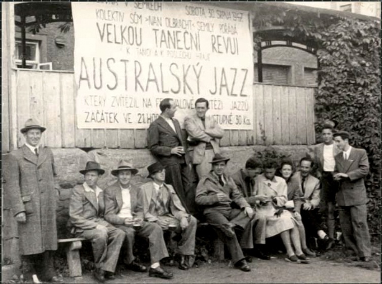 Group of men in long coats and hats, with two women, standing and seated outside in front of a fence. Above them is a large white banner with black foreign text. Behind the fence and the banner is a brick building.