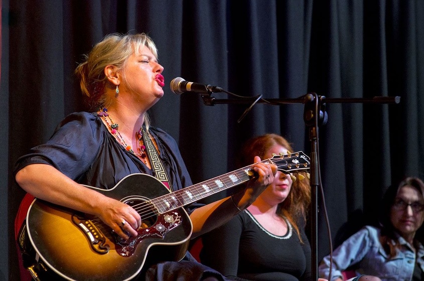 Colour photograph of a seated woman wearing a black top playing a guitar and singing into a microphone. Behind are two other women, and a black curtain behind the three of them.