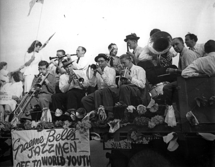 Black and white photograph of a group of men seated on a float playing instruments including a trombone, three trumpets, two clarinets andand a tuba.. Flowers and a banner is beneath their feet hanging from the side of the float reading 'Graeme Bells Jazzmen off to world youth"