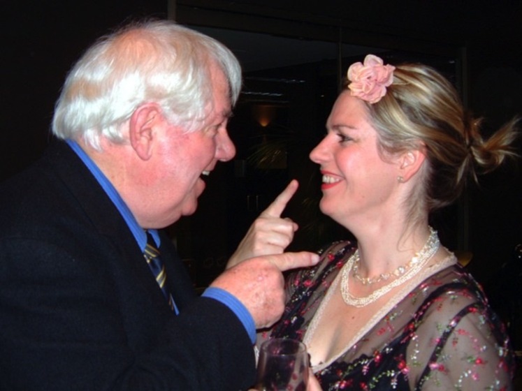 Colour photograph of a man in a suit and blue shirt with a tie and grey hair, pointing at a woman in front of him. The women faces him ponting at her own nose, and wears a black top with a net overlay with pink flowers, and a pink flower in her hair.  