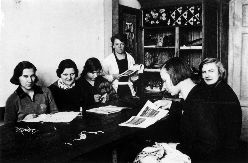 Black and white photograph of a group of women seated around a table, with magazines. Behind them is a set of wooden shelves.