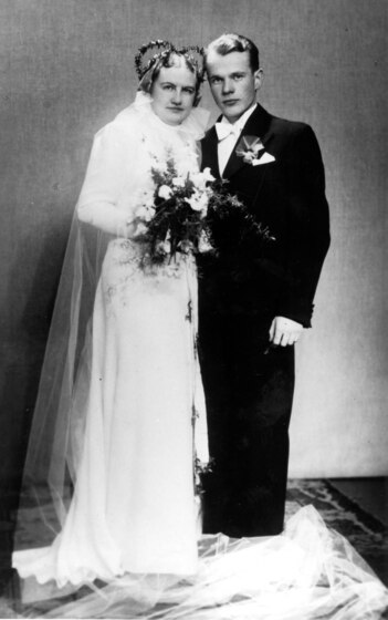 Black and white studio photograph of a man and a woman. The woman wears a white wedding dress and holds a bouquet of flowers. The man wears a suit and has a handkerchief in in breast pocket. 