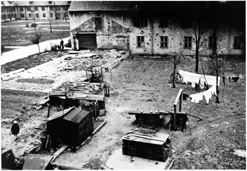 Black and white photograph of a dirt yard with four shacks and two washing lines of white clothes, in front of a long two-story building. There are a small number of thin trees with no leaves across the ground. 