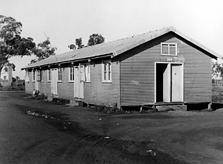 Black and white photograph of a weatherboard building sitting on concrete blocks, on a dirt surface.