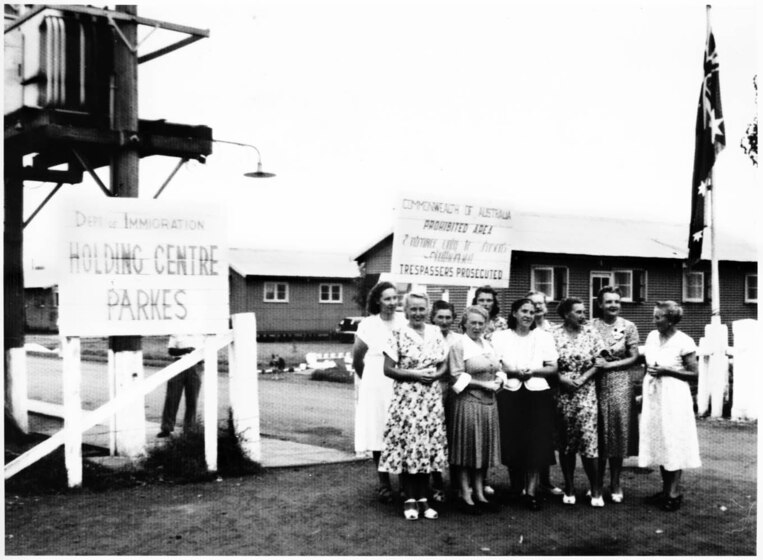 Black and white photograph of a group of women standing together in front of a weatherboard building. There is a sign to their left reading 'Holding Centre Parkes', and a flagpole to their left. Behind them is another sign reading 'Commonwealth of Australia Prohibited Area Trespasers Prosecuted'.