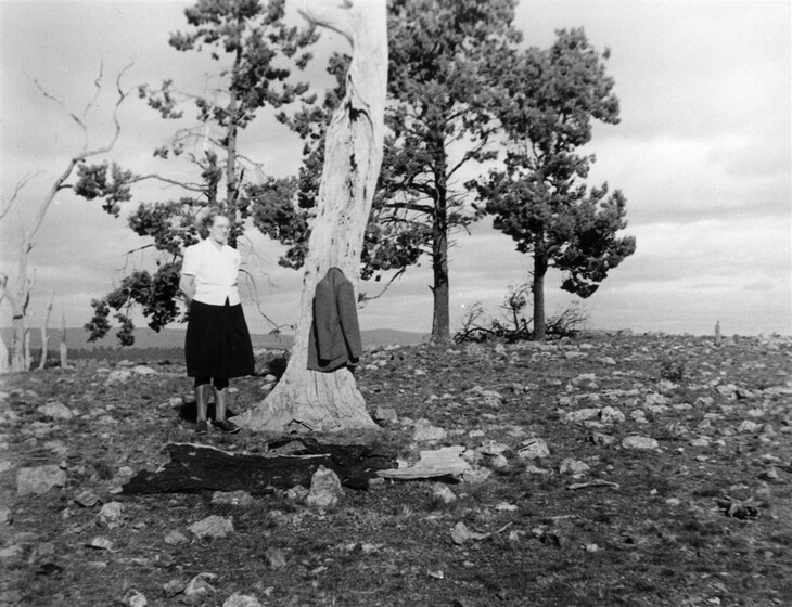 Black and white photograph of a woman standing next to a tree in a rocky field. There is a jacket hanging on the tree trunk.