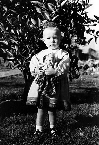 Black and white photograph of a young child holding a doll, standing in front of a bush.