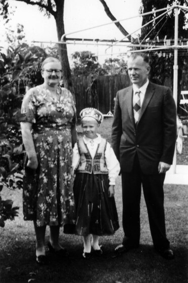 Black and white photograph of a young girl is a long skirt and headdress, standing between a woman on the left in a floral dress, and a man on the right in a suit. There is a Hill Hoist clothesline behind.