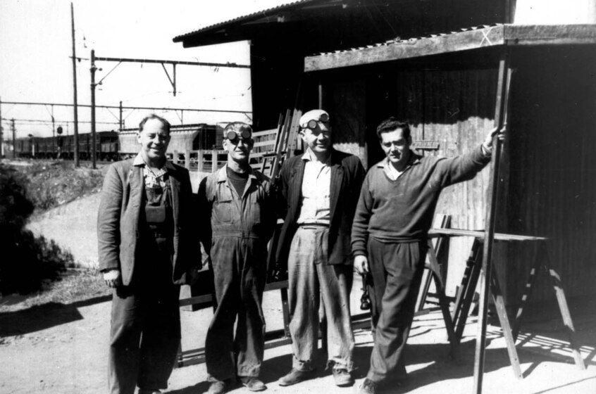 Black and white photograph of four men in work wear standing in front of a shack. There is a train seen in the distance behind.