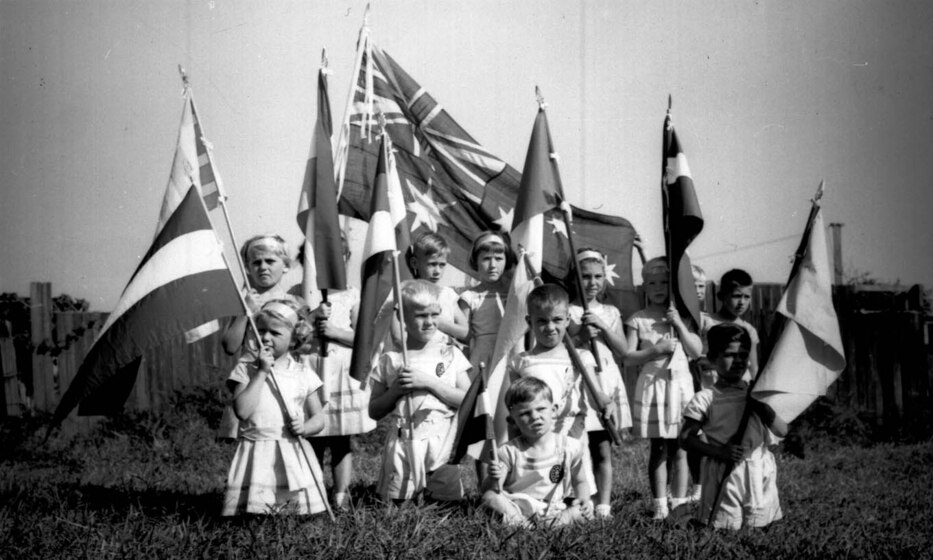 Black and white photograph of a group of children sitting on grass, holding a range of flags on sticks from different nations.