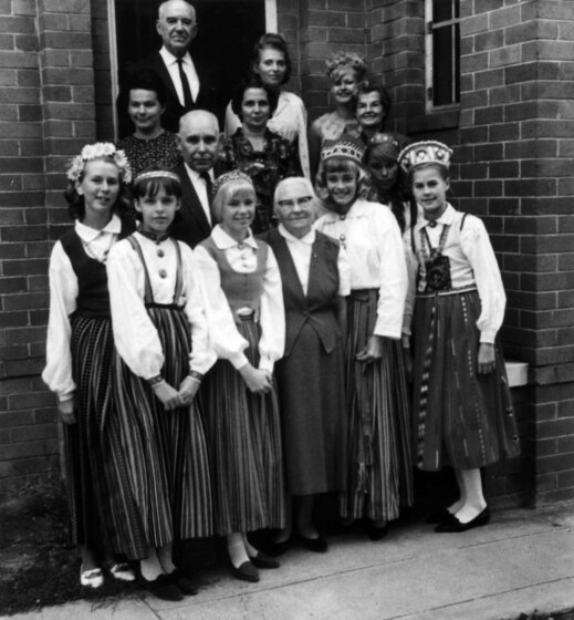 Black and white photograph of a group of young girls in long skirts and headdresses, standing in front of a brick building with eight other men and women.
