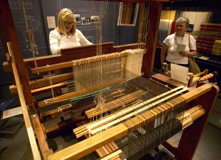 Colour image of a large loom with white and black thread wound on it. Behind stands two women.