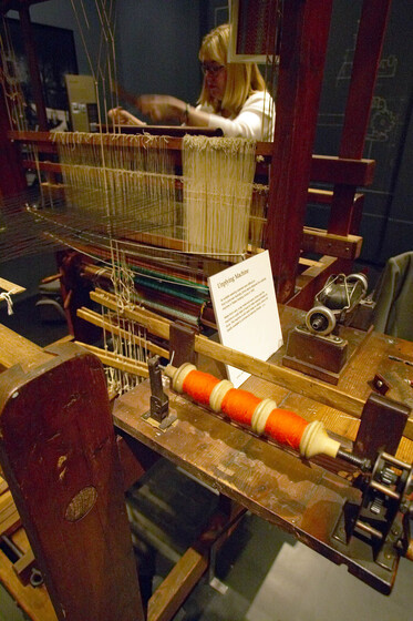 Colour image of a large loom, showing a working table with a white card with text on it, and a spool with orange thread. There is a woman standing behind the loom. 