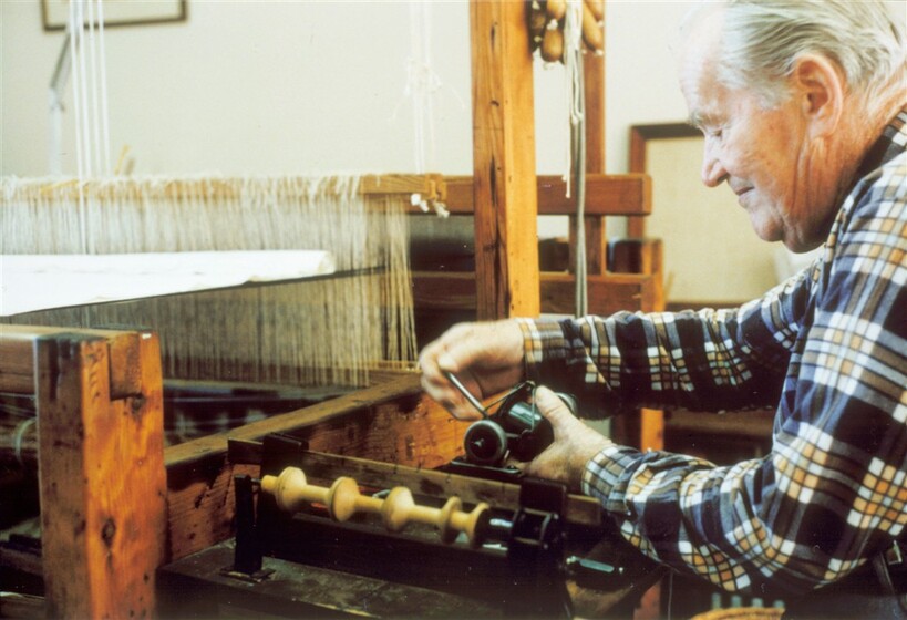 Colour photogtraph of an older man in a blue, white and yellow check shirt, seated at a large loom, holding a piece of the equipment.