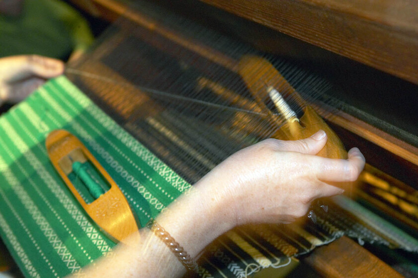 Close up photograph of two boat shuttled being used on a large loom. One shuttle sits on a woven green and white fabric.