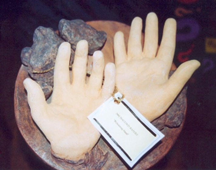 Two ceramic hands on rocks, on a circular piece of timber. On the two hands is a small white card with unreadable text.