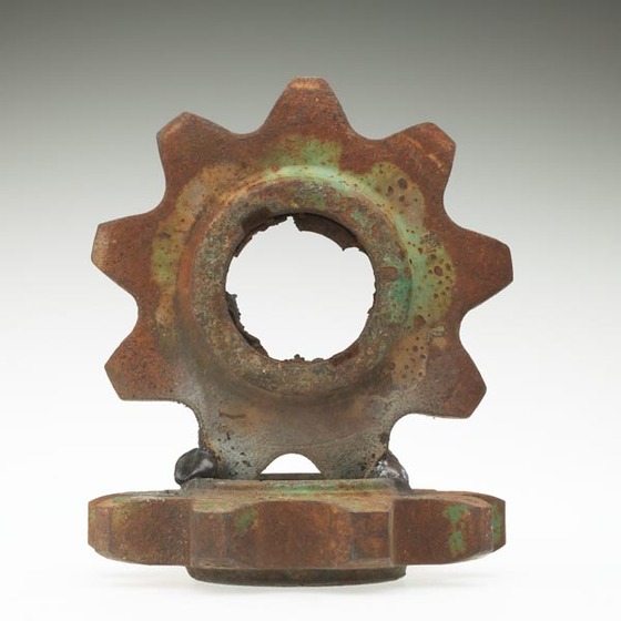Two large metal cogs. One cog is laying down, the other is welded to the top standing upright. 