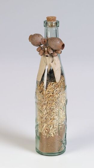 Glass bottle containing sand and seeds. It is corked, and has gum nuts and gum leaves tied around the neck.