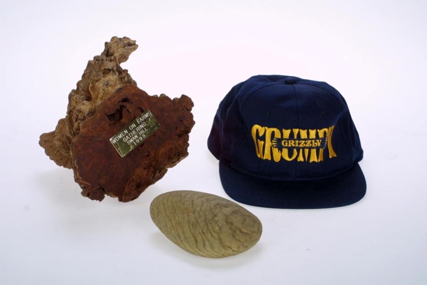 A piece of timber with a small gold plaque fixed to it, a smooth stone, and a blue peaked cap with yellow words reading 'grumpy grizzly' on the front.