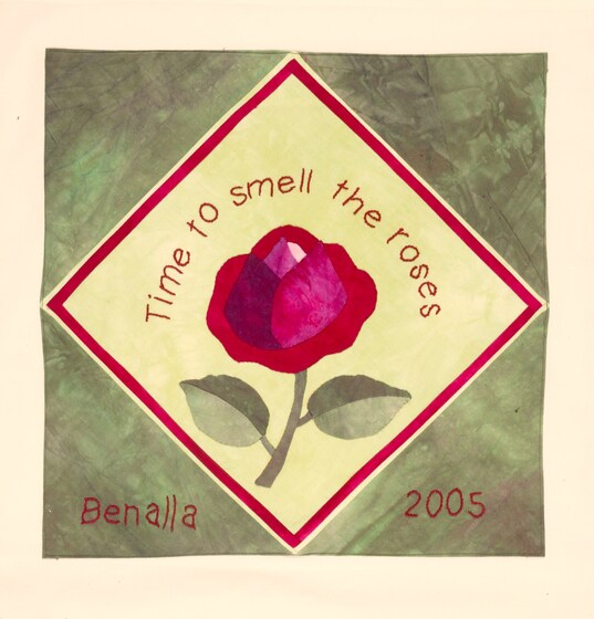 Fabric square with a rose in the middle of a yellow diamond on a green background. Red text above the rose reads 'time to smell the roses'. Beneath the diamond in red text reads 'Benalla 2005'. 