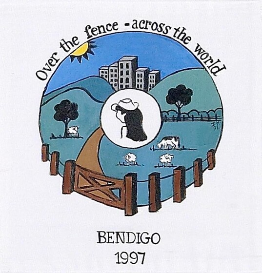 White fabric square with a circular colour image of a farm with city buildings in the distance. In the centre is a white circle with a women's head. Beneath is the text 'Bendigo 1997'. Above is the text 'Over the fence - across the world'. 