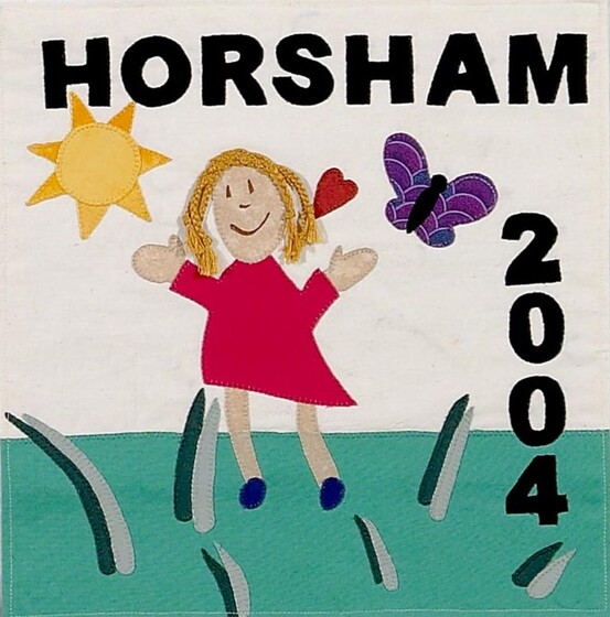 White fabric square of a young girl in a red dress standing in a grass field with a sun and purple butterfly above. Black text reads 'Horsham 2004'. 