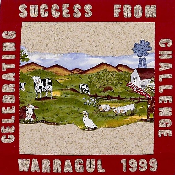 Red fabric square with a farm scene featuring cows, sheep and ducks on rolling green hills in the centre. Surrounding in white text on the red reads 'celebrating success from challenge, Warragul 1999'. 