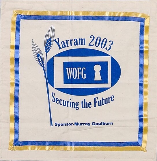 White fabric square with a blue and yellow ribbon square border. In the centre is a blue logo of wheat with an oval and the acronym 'WOFG' in the middle next to a keyhole. Above is text 'Yarram 2003'. Below is text 'Securing the Future'. 