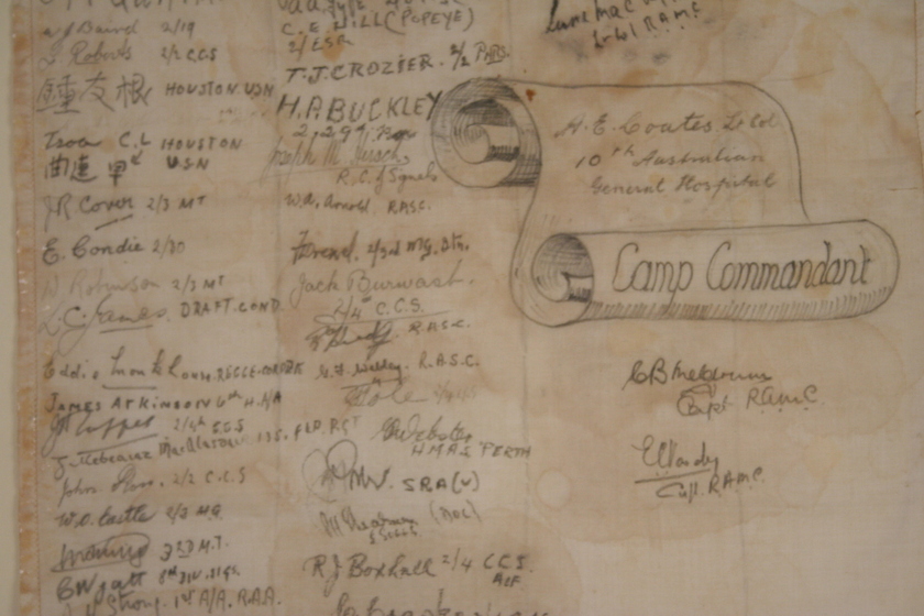 Close up view of paper that is yellow and brown with age with three columns of handwritten names. A hand drawn paper scroll with the title 'Camp Commandant' written on the roll is on the right. 