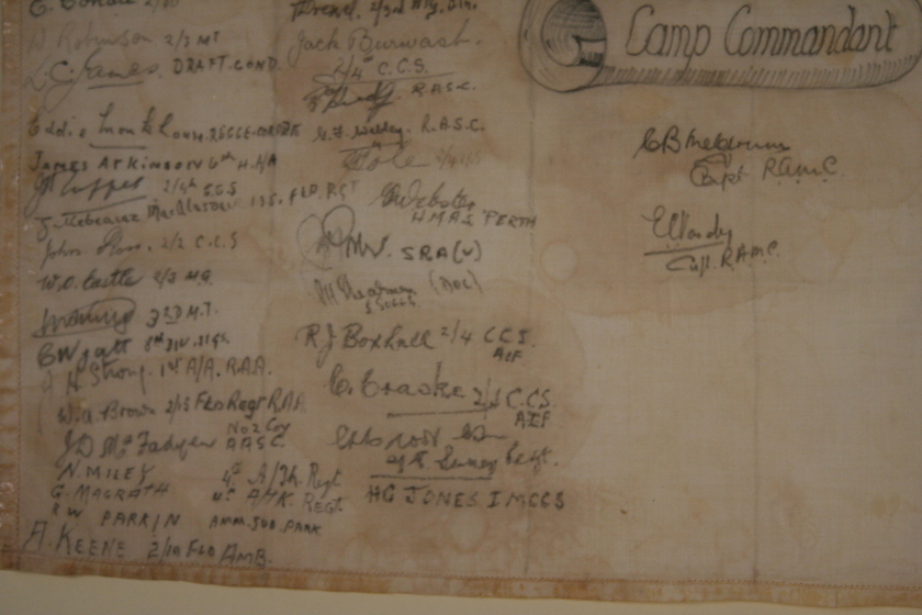 Close up view of paper that is yellow and brown with age with three columns of handwritten names. The bottom of a hand drawn paper scroll with the title 'Camp Commandant' written on the roll is on the top right