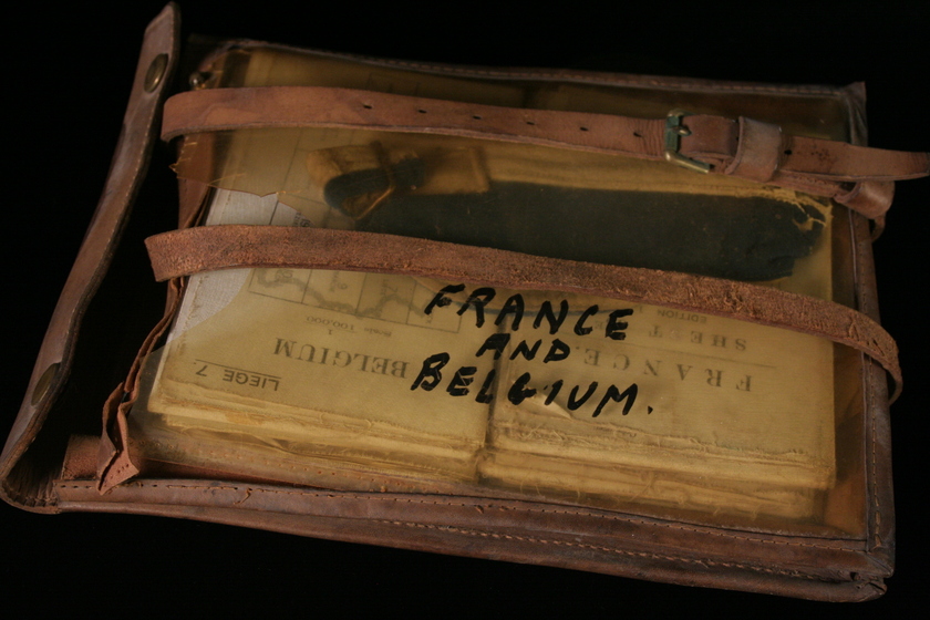 Leather satchel with one transparent yellowed side, containing a stack of folded maps. Two leather straps and buckles laying over the top. In marker written on the transparent side is the text 'France and Belgium'. 