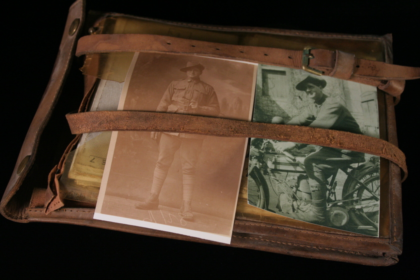 Leather satchel with two black and white photographs of men in army uniforms, sitting on top of one transparent yellowed side. Two leather straps and buckles laying over the top of the satchel and photographs.