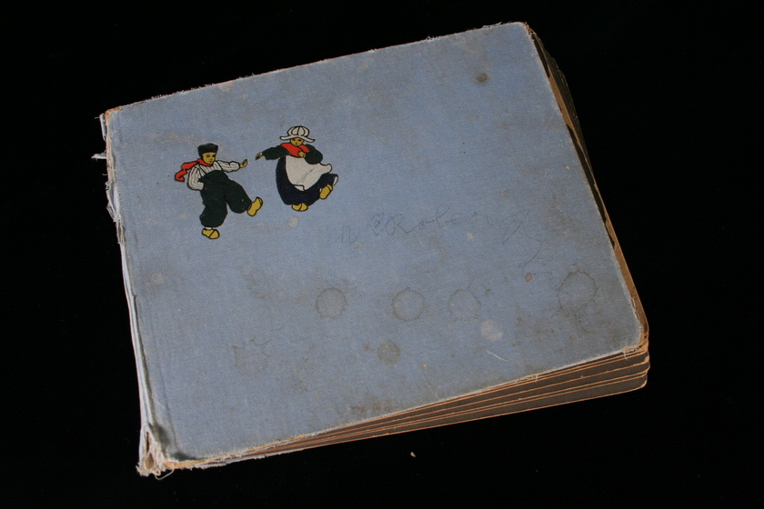 View of the front cover of a thick light coloured photo album on a black background, with two cartoon figures of a man and a woman in black and white clothing with red scarfs in the top left corner. The esdges of the album show fraying. 