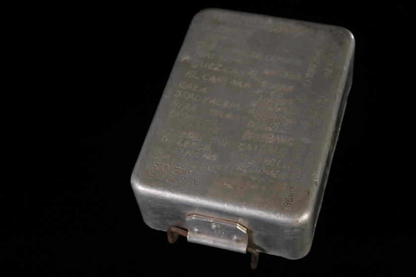 Metal rectangular box viewed from above, with a hinge on the bottom front side. There is writing engraved across the entire top of the box.
