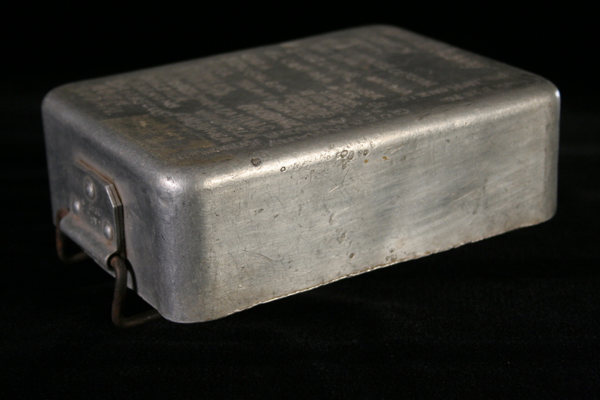 Metal rectangular box viewed from the side, with a hinge on the bottom front side. There is writing engraved across the entire top of the box.