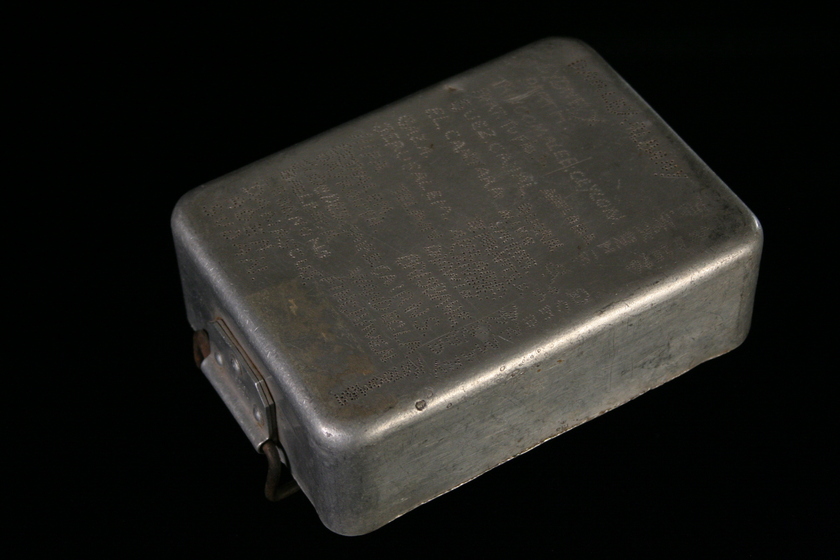 Metal rectangular box viewed from above, with a hinge on the bottom front side. There is writing engraved across the entire top of the box.