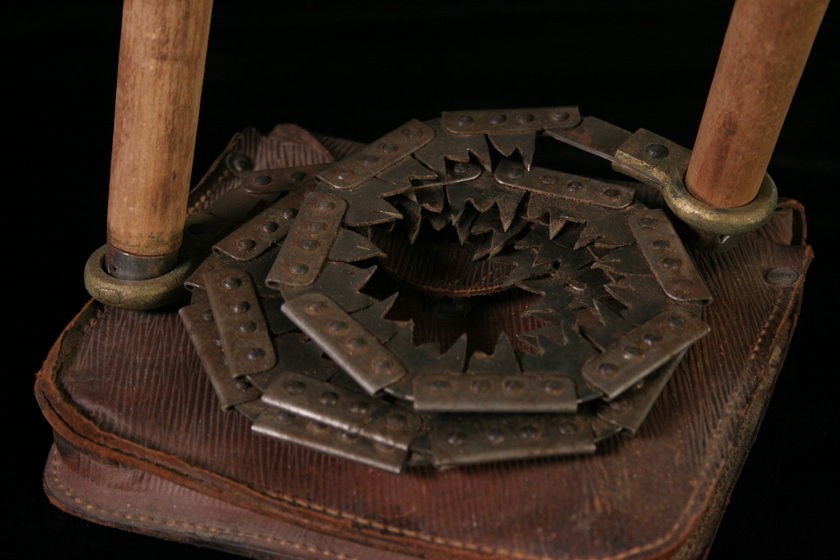 Close up view of a coiled hand-held saw, with two wooden handles, and the teeth of the metal saw coiled around to form circles. The saw is sitting on a leather pouch.