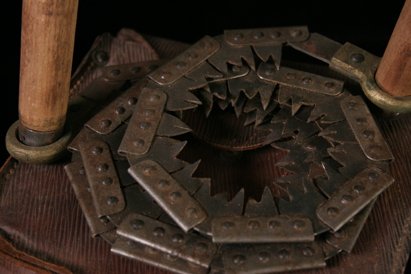 Close up view of a coiled hand-held saw, with two wooden handles, and the teeth of the metal saw coiled around to form circles. The saw is sitting on a leather pouch.