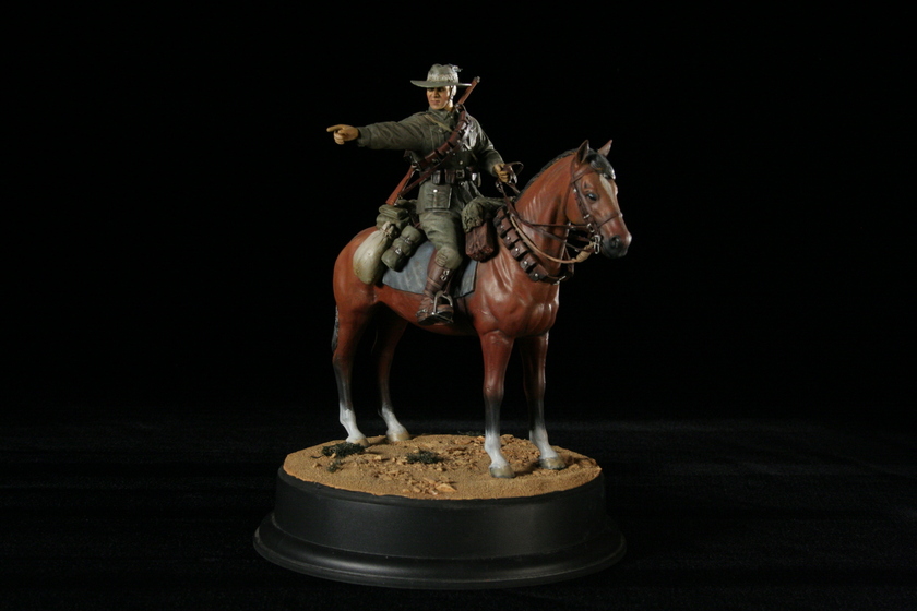 Close up view of a figurine of a man seated on a brown horse in a military army uniform, viewed from the right side of the horse. The man is pointing to the left.